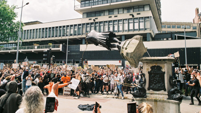 In a town centre, a crowd watches as a bronze statue is pulled from its high stone plinth by ropes