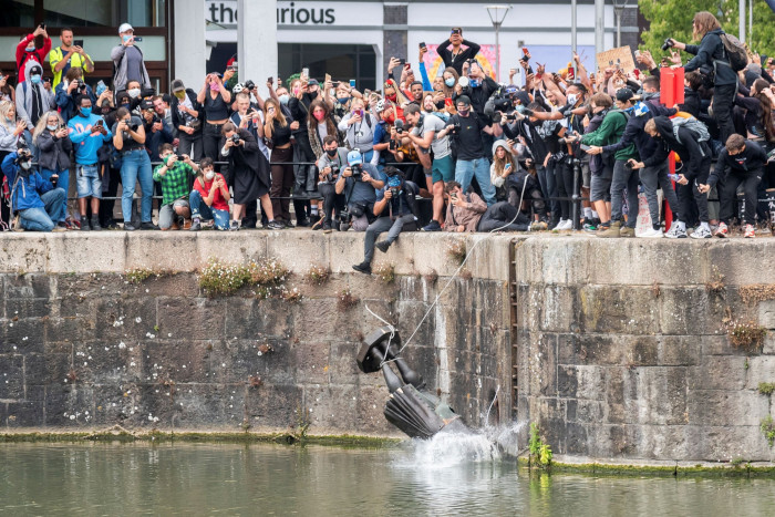 A crowd gathers along a quay, cheering as the bronze statue hits the water head first, a rope still attached around its ankles