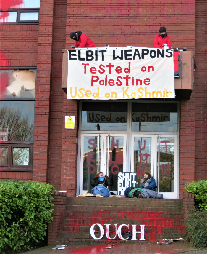Entrance to a red-brick building. Two people are in freont of the door in sleeping bags. Above the doorway is a balcony, on which two more people stand. They are fixing a banner reading: Elbit Weapons tested on Palestine, used on Kashmir. Red paint has been poured over windows and steps.