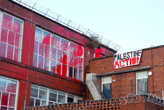 1930s-style factory building with razor wire draped along the edges. The tall windows have been splashed with red paint and a banner reads Palastine Action.