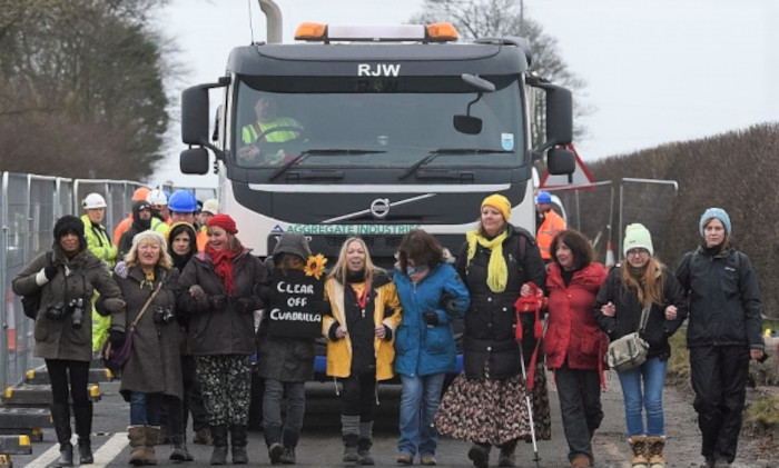 A lane, hemmed in with haras fencing, with a lorry. In front of the lorry a group of about 11 people are walking, arm in arm. Behind the people are a few people in hard hats and hi-viz jackets.