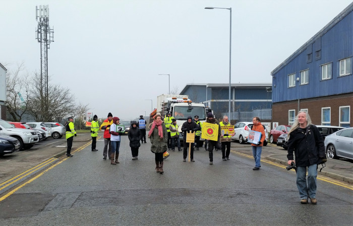 A road on an industrial estate, units on one side, cars parked on the other. A small group of people are walking towards the camera, most with a placard in their hands. At the back of the photograph, between the protestors and a lorry are a number of police officers.