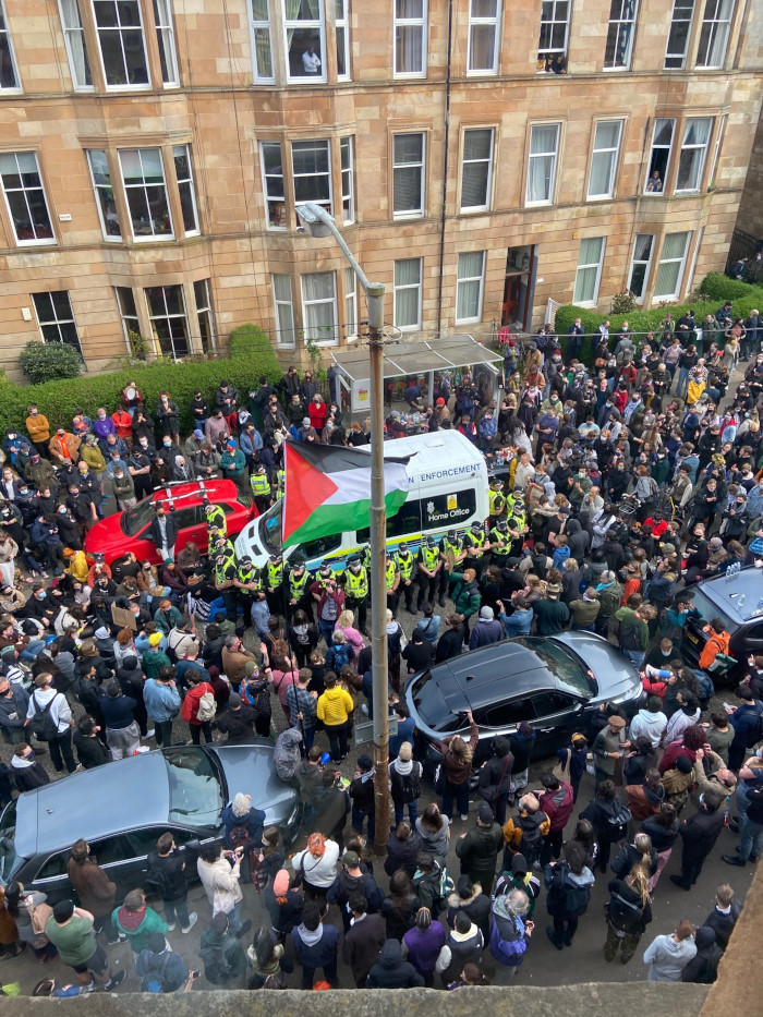Seen from above, a narrow street choked with people. In the centre of a crowd, an immigration van is surrounded by a single line of police officers in hi-viz vests.