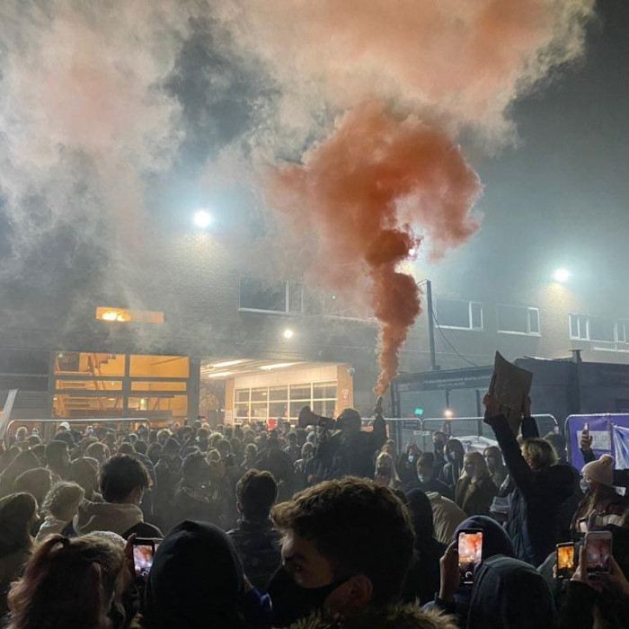 It's dark, a crowd of people in front of an institutional-looking building. A silhouetted figure in the middle holds a loudhailer and an orange flare.