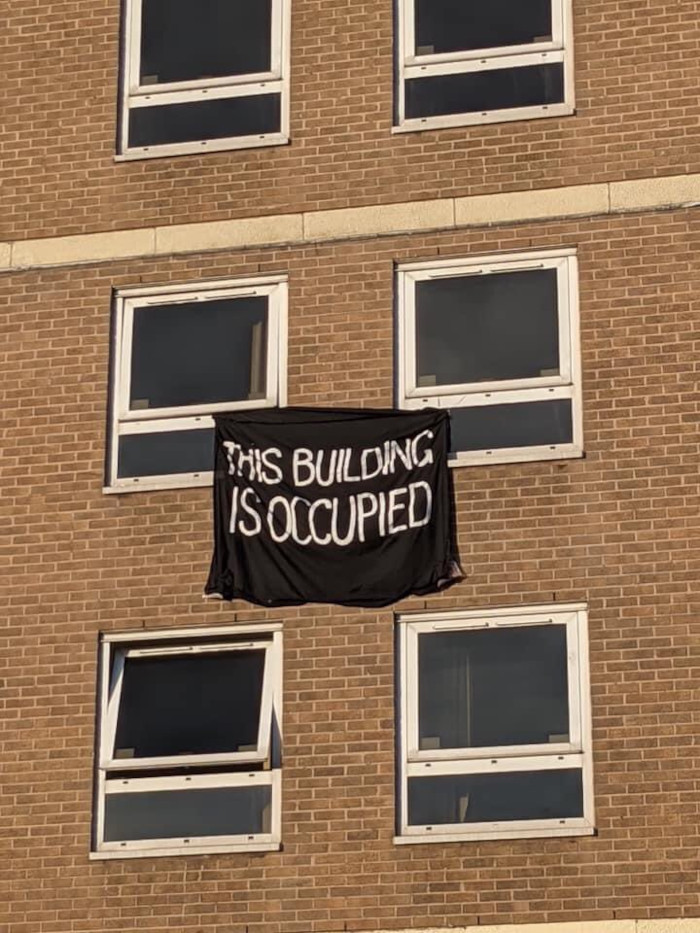 A yellow brick building with a banner hanging from windows reading: This Building Is Occupied